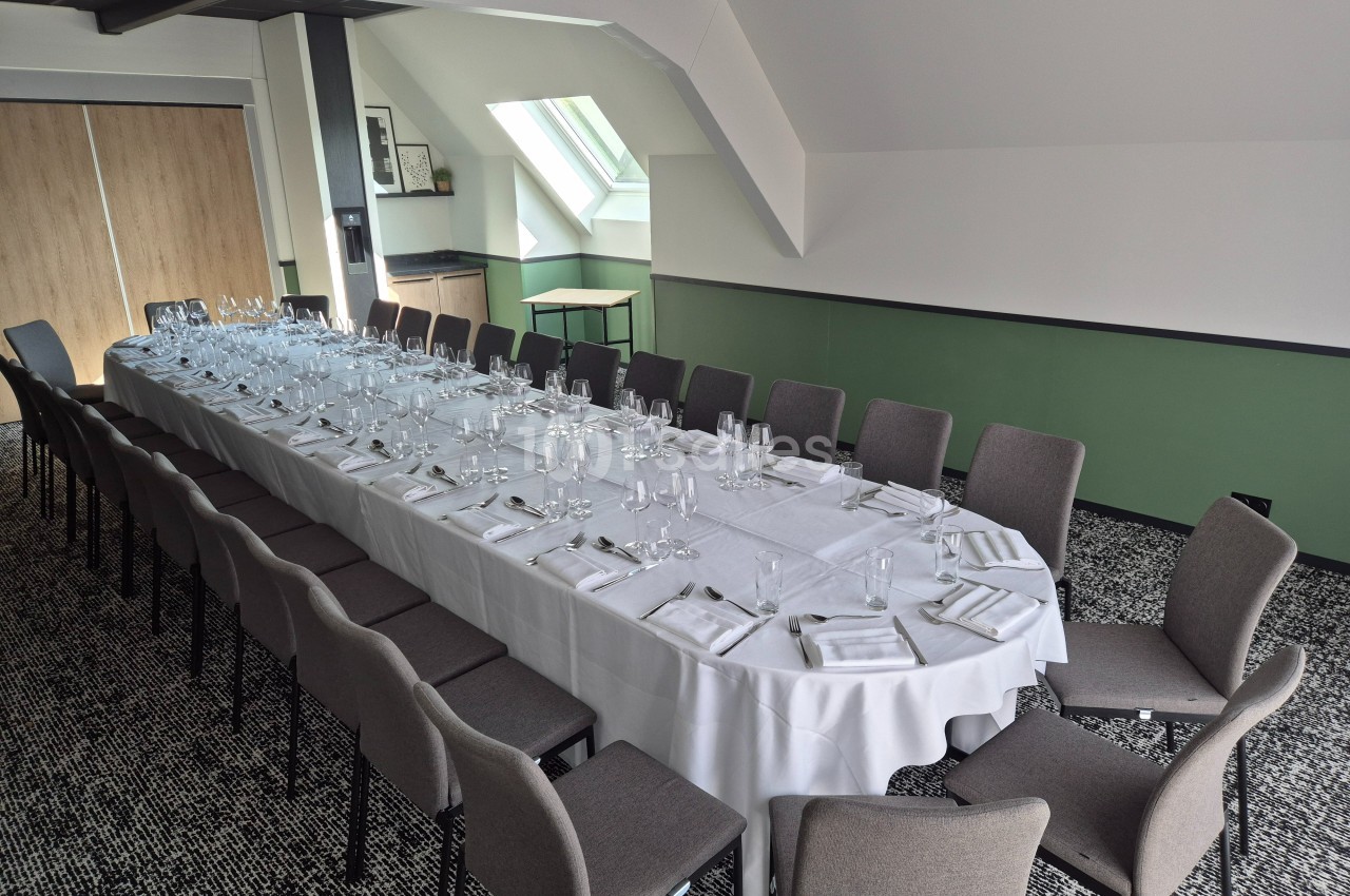 Salle de réception avec une grande table dressée, entourée de chaises, sous un plafond mansardé.