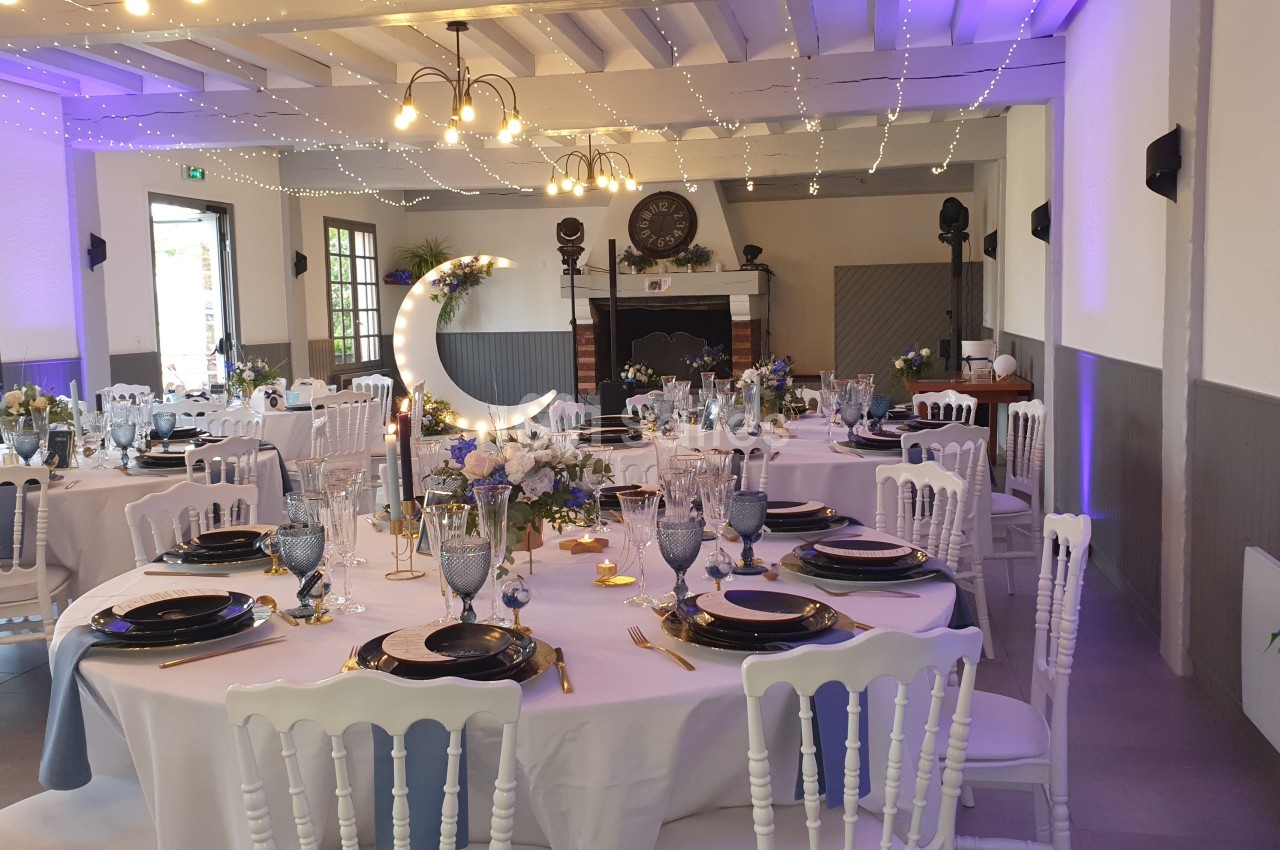 Salle de réception décorée avec des tables rondes dressées, éclairage tamisé et guirlandes lumineuses suspendues.