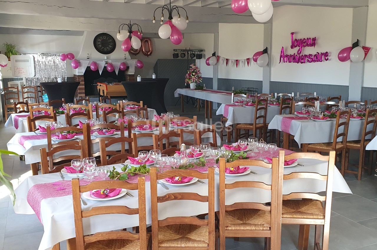 Salle décorée pour un anniversaire avec des tables dressées, des ballons roses et blancs, et une banderole ’Joyeux…