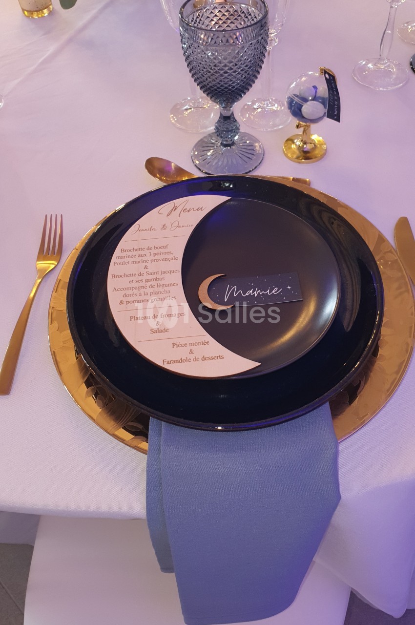 Assiette noire avec un menu imprimé et une serviette bleue, disposées sur une table dressée avec des couverts dorés.