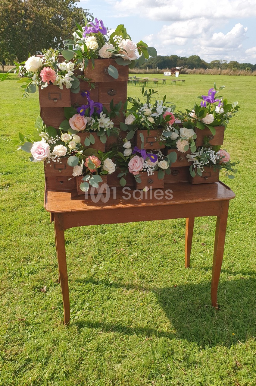 Arrangement floral dans des tiroirs en bois empilés sur une table en bois, posé sur une pelouse sous un ciel dégagé.