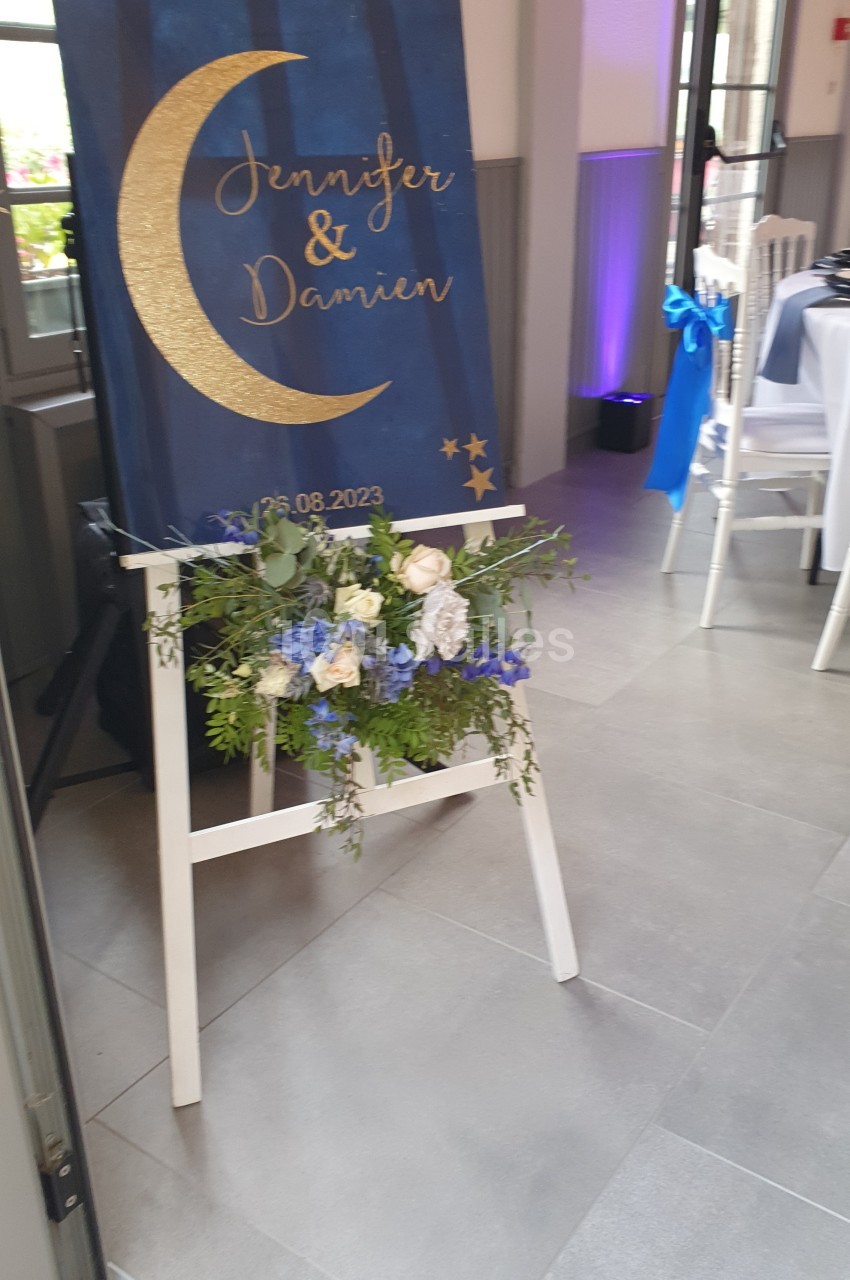Panneau de bienvenue avec des noms et une date, décoré de fleurs bleues et blanches, placé sur un chevalet.