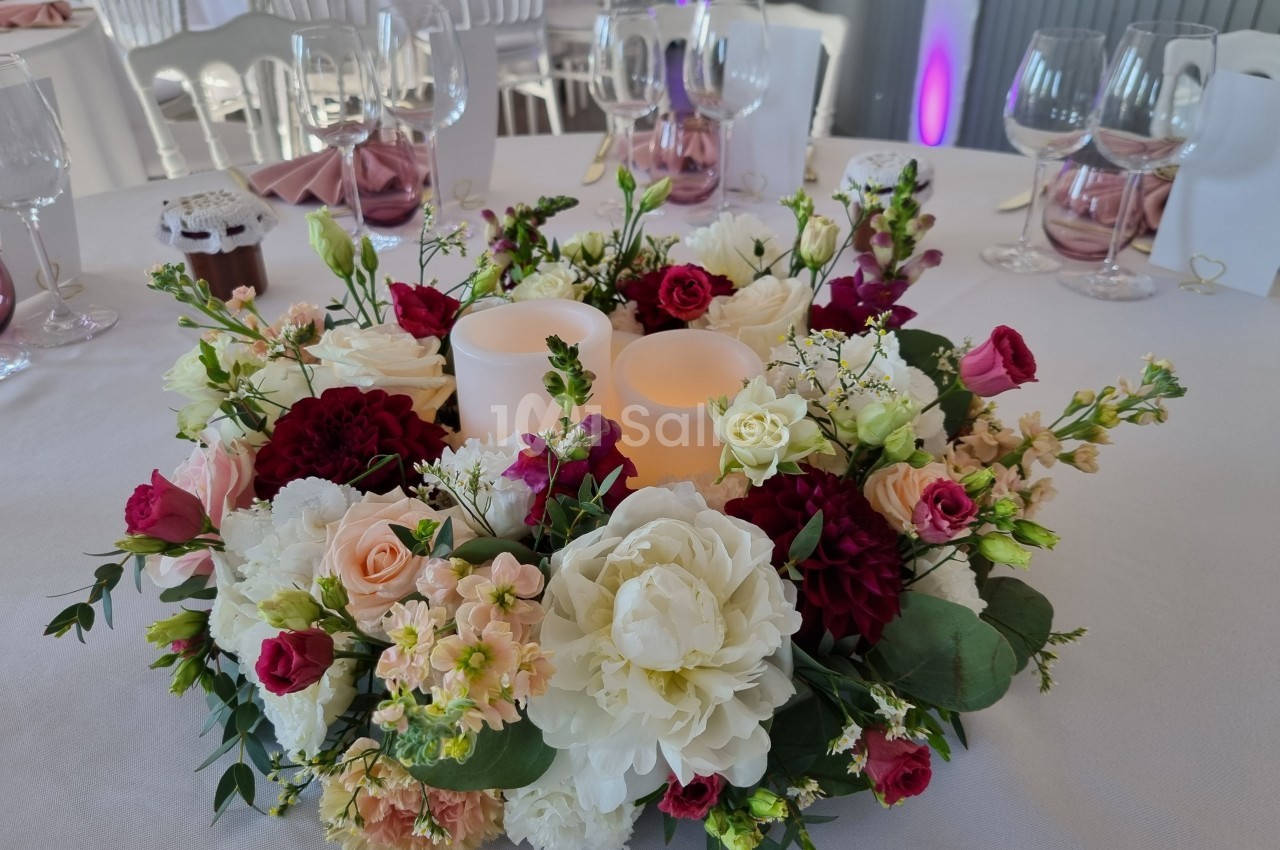 Centre de table floral avec bougies LED entourées de roses, pivoines et feuillage, sur une nappe blanche.