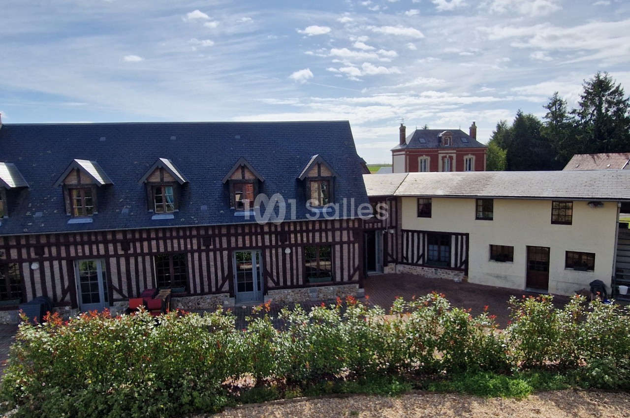 Bâtiments en colombages et en briques avec toiture en ardoise, entourés de végétation sous un ciel partiellement nuageux.
