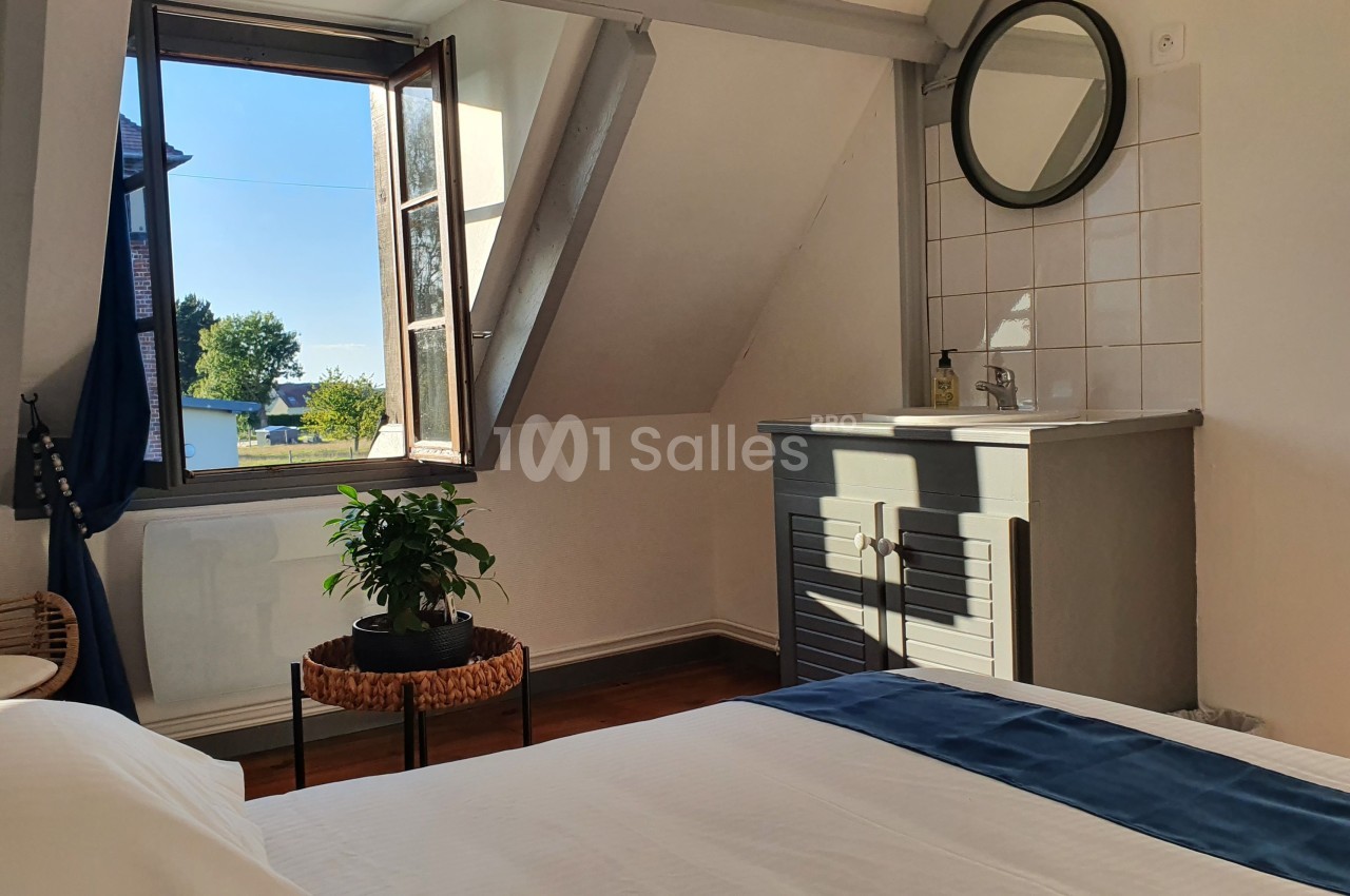 Chambre lumineuse avec lit, fenêtre ouverte sur un paysage, lavabo avec miroir et plante sur une table basse.