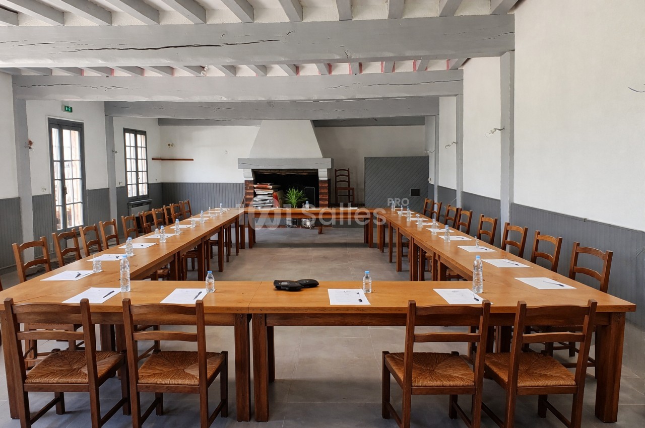 Salle de réunion avec tables disposées en U, chaises en bois, bouteilles d'eau et documents posés sur les tables.