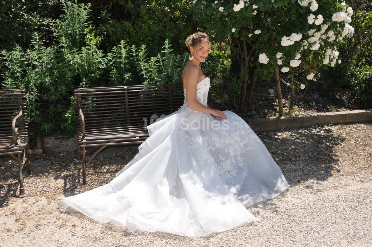 Femme en robe de mariée blanche assise sur un banc en métal, entourée de verdure et de fleurs blanches.