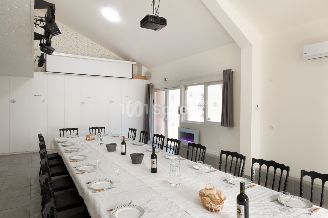 Salle lumineuse avec une grande table dressée pour un repas, chaises noires et projecteur au plafond.
