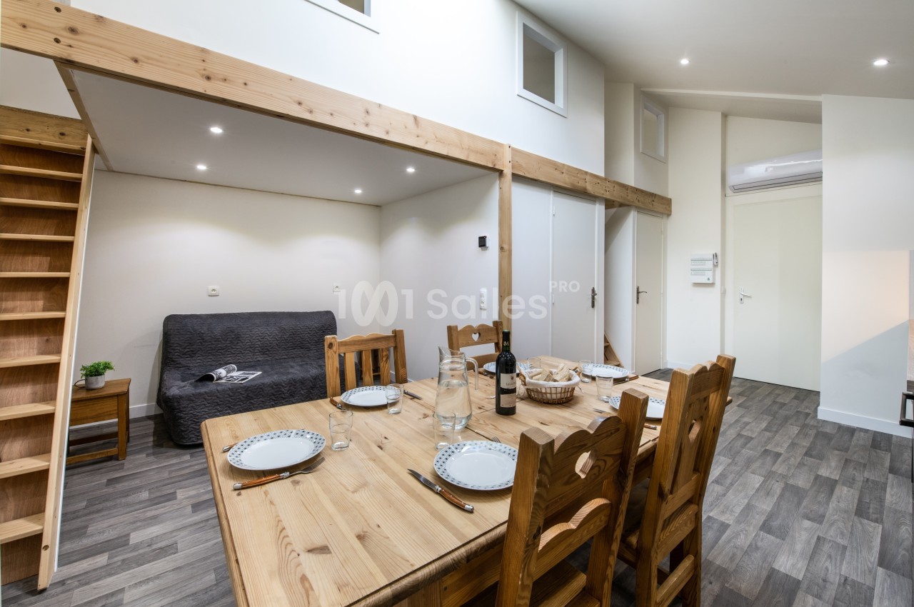 Salle à manger avec table en bois dressée, canapé gris en arrière-plan et mezzanine visible au-dessus.