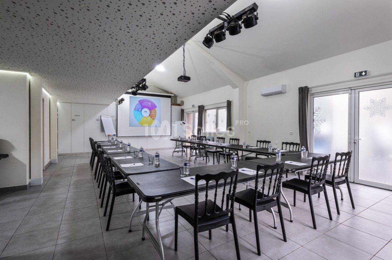 Salle de réunion lumineuse avec tables disposées en U, écran de projection allumé et chaises noires.