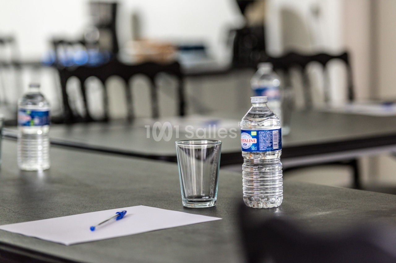 Bouteille d'eau, verre vide, stylo et feuille posés sur une table dans une salle avec des chaises en arrière-plan.