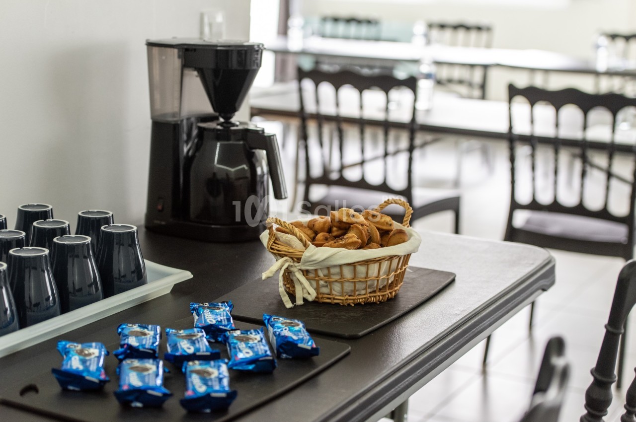 Table avec cafetière, panier de biscuits, gobelets noirs et snacks emballés dans une salle avec chaises en arrière-plan.