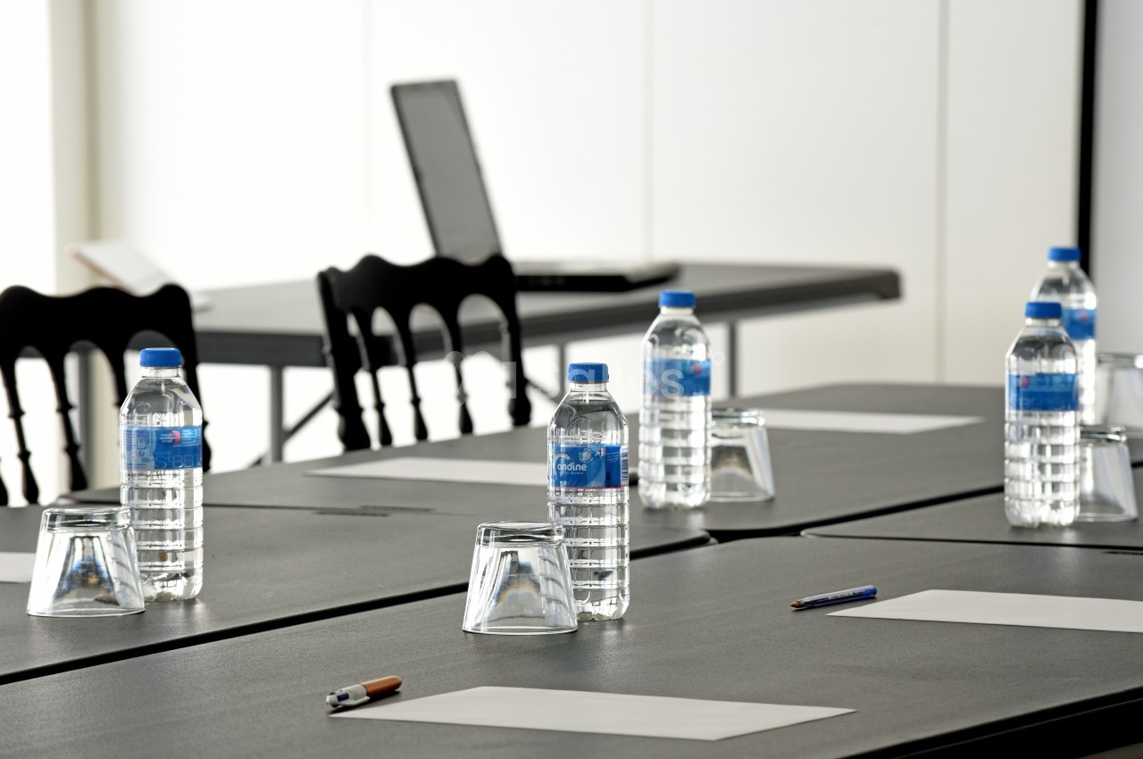 Salle de réunion avec tables noires, bouteilles d'eau, verres, stylos et feuilles, ordinateur en arrière-plan.