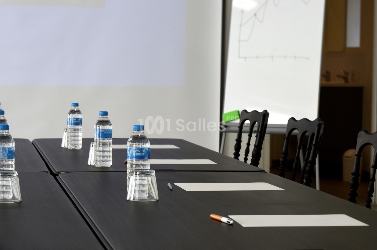 Salle de réunion avec table noire, bouteilles d'eau, feuilles blanches, stylos et tableau blanc en arrière-plan.