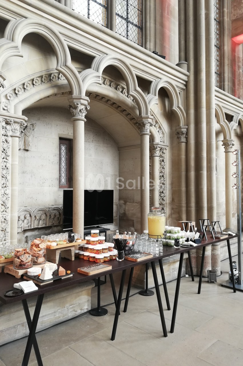 Buffet disposé sur des tables dans un espace architectural avec arches en pierre et vitraux en arrière-plan.