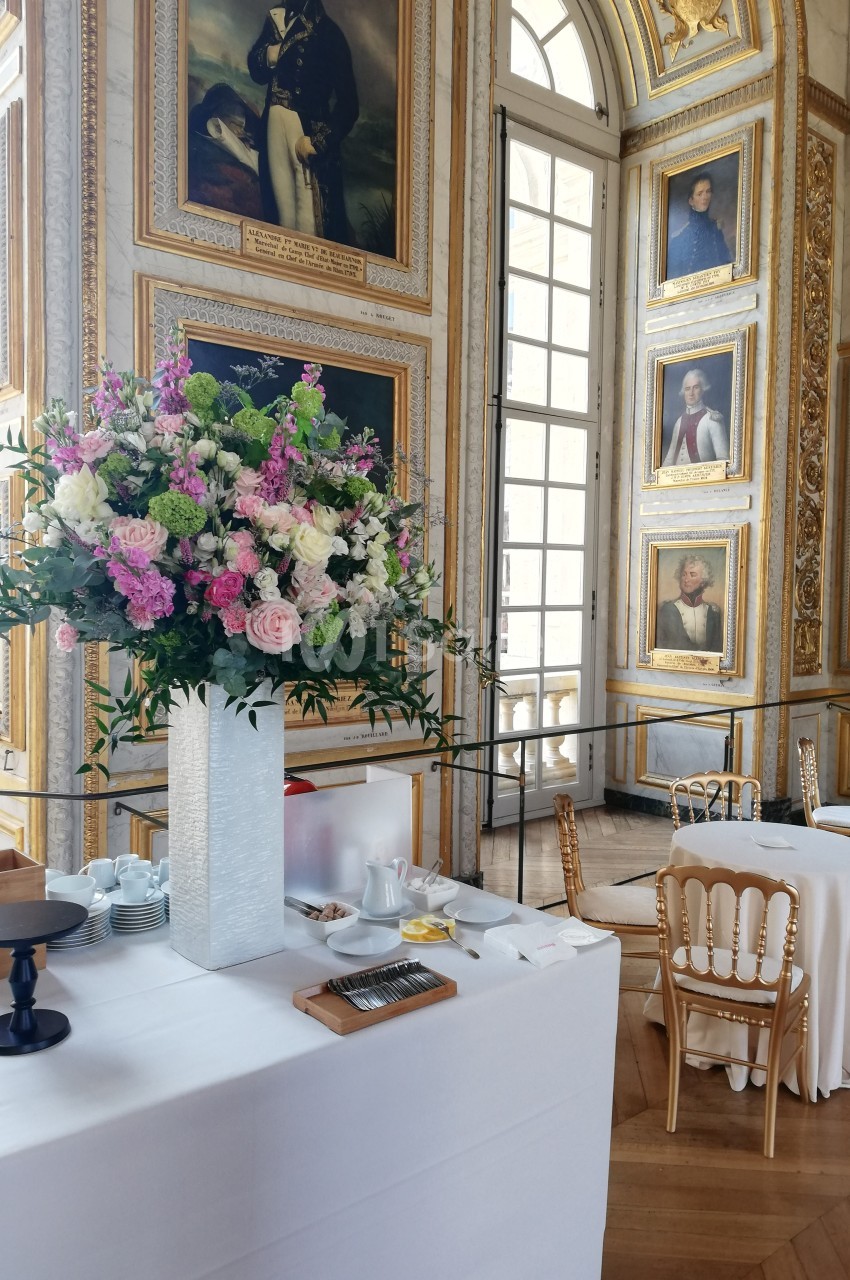 Grande salle ornée de portraits et dorures, avec une table dressée, décorée d'un grand bouquet de fleurs colorées.