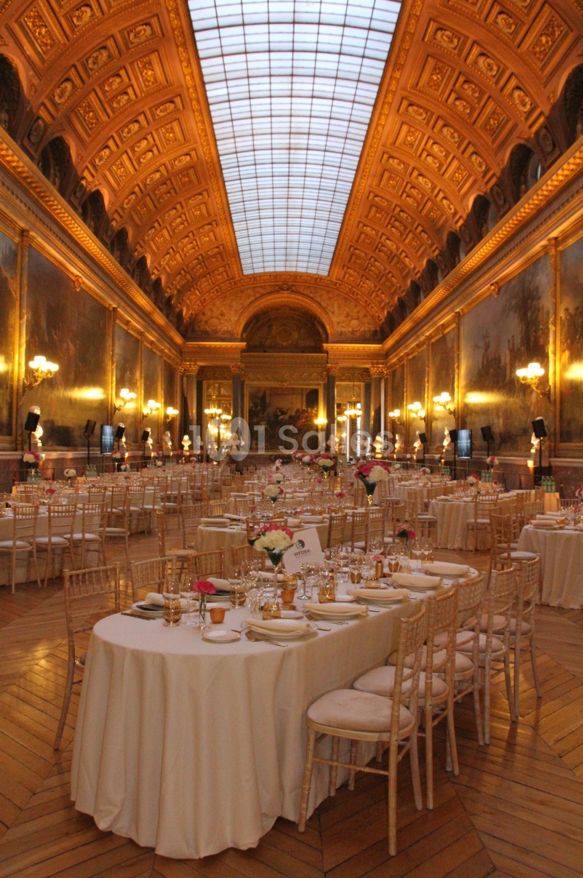 Salle de réception élégante avec tables rondes dressées, éclairée par des lustres et un plafond en verrière.