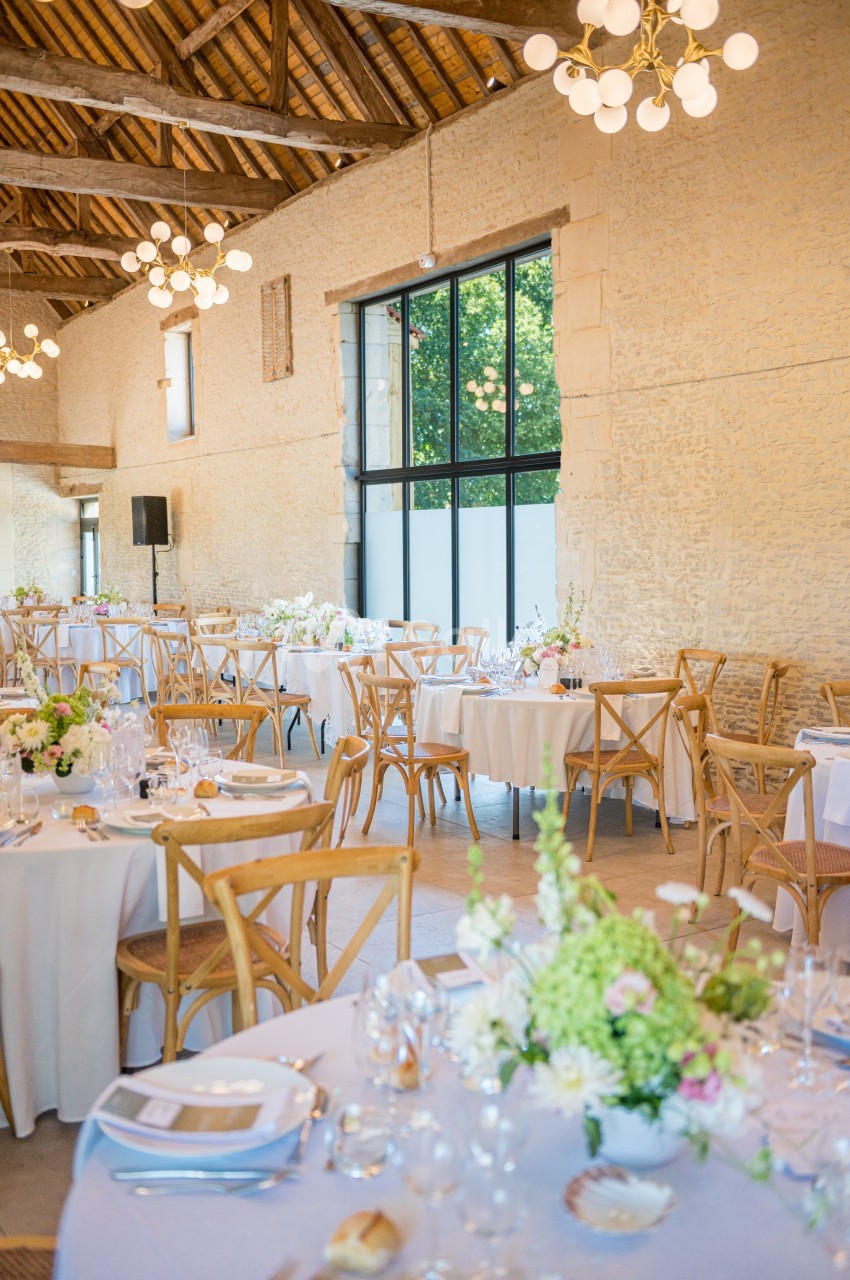 Salle de réception lumineuse avec tables décorées de fleurs, chaises en bois et grandes fenêtres donnant sur l'extérieur.