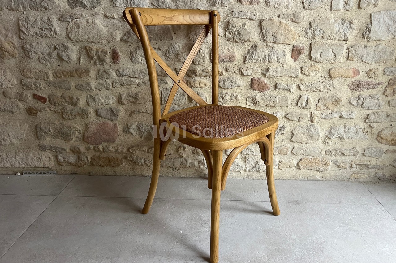 Chaise en bois clair avec dossier croisé et assise en rotin, posée sur un sol en carrelage devant un mur en pierre.