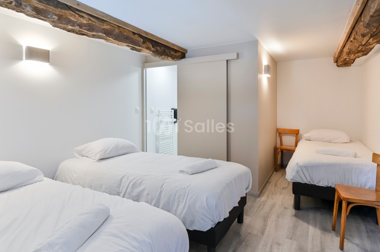 Chambre lumineuse avec trois lits simples, murs blancs, poutres apparentes et sol en parquet clair.