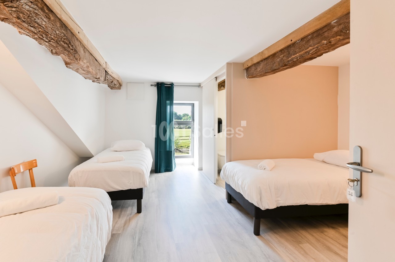 Chambre lumineuse avec trois lits simples, poutres apparentes et vue sur l'extérieur par une porte vitrée.