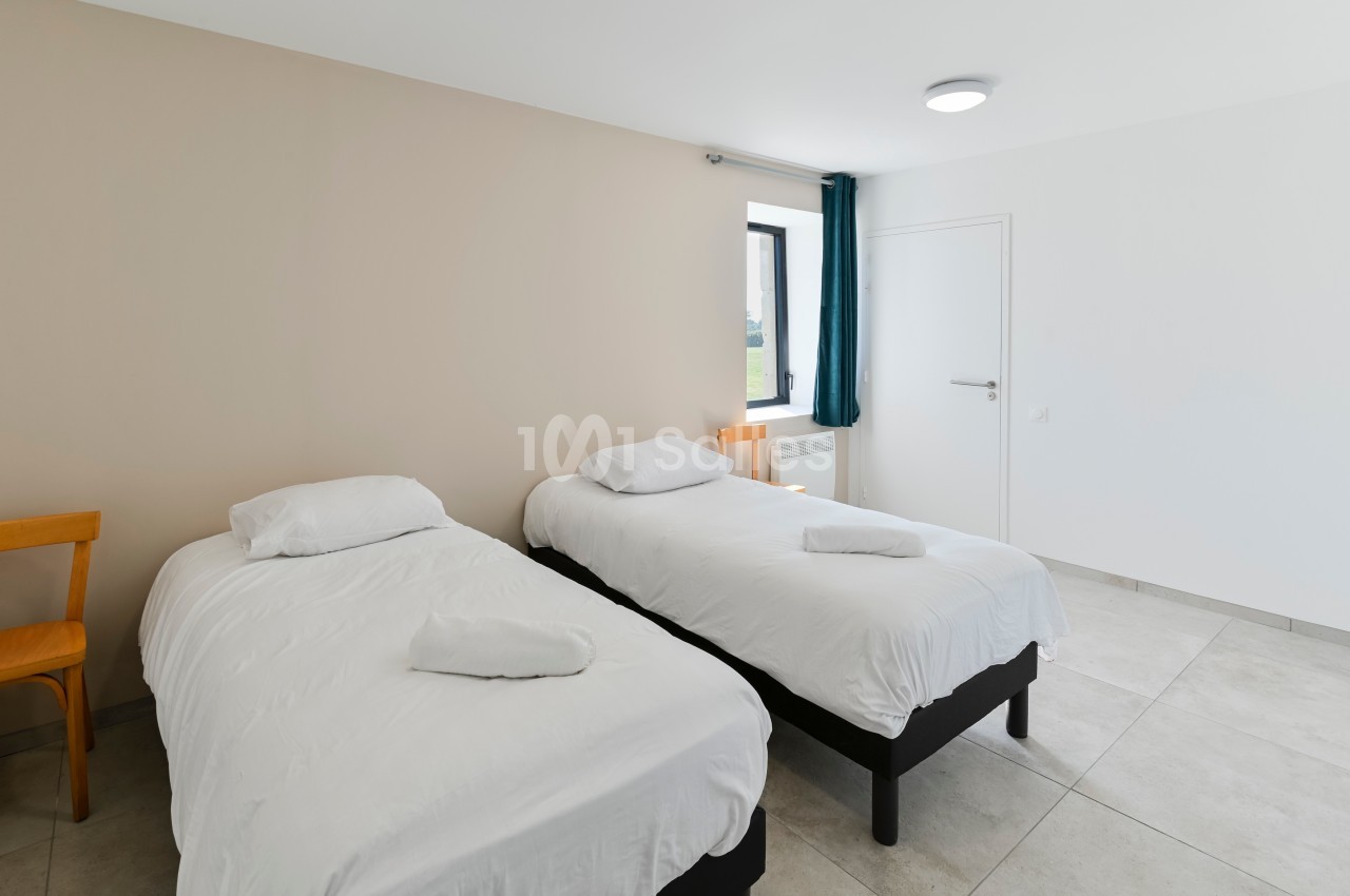 Deux lits simples avec draps blancs dans une chambre lumineuse, équipée d'une chaise en bois et d'une fenêtre.
