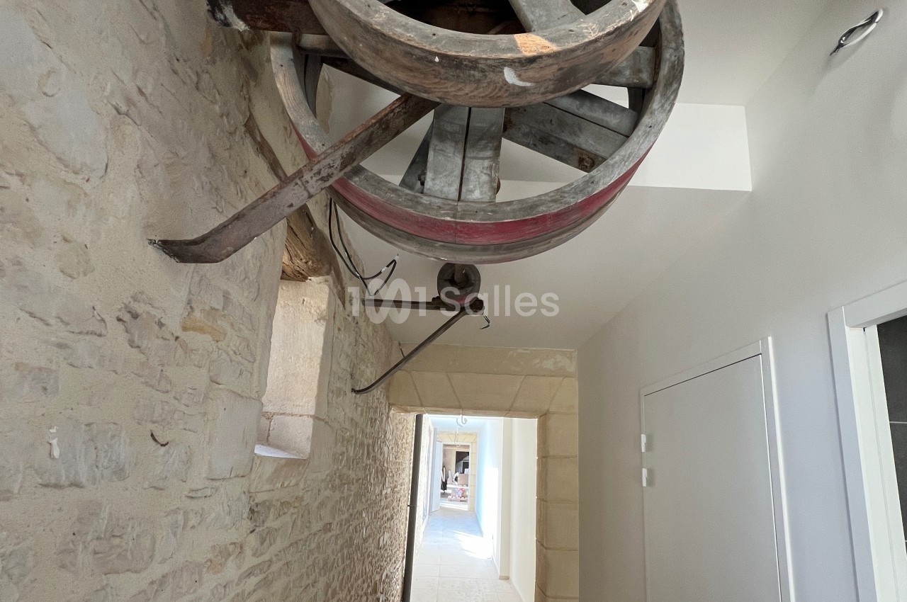 Roue en bois suspendue au plafond d'un couloir aux murs en pierre, avec une porte et un passage lumineux au fond.