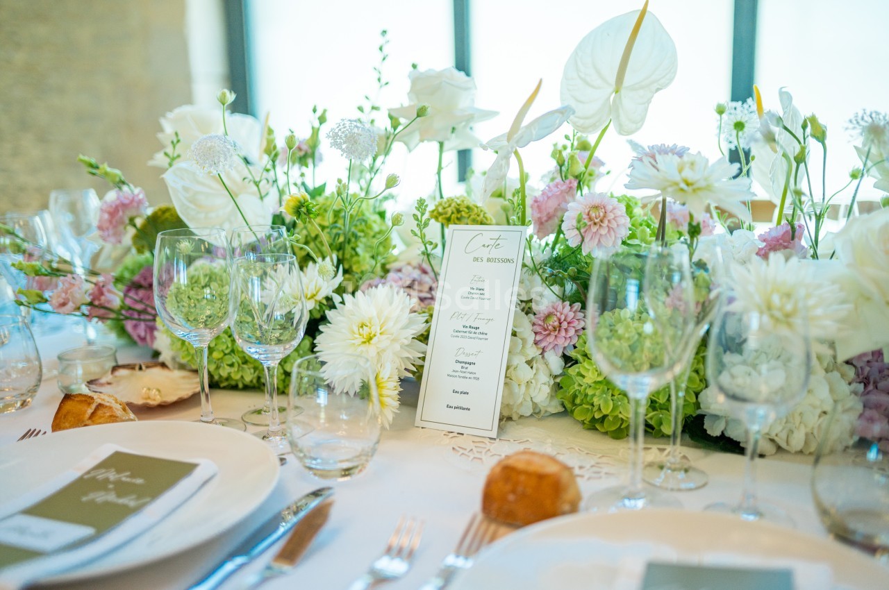 Table élégamment dressée avec des assiettes, verres, menus et un centre de table floral aux tons blancs et verts.
