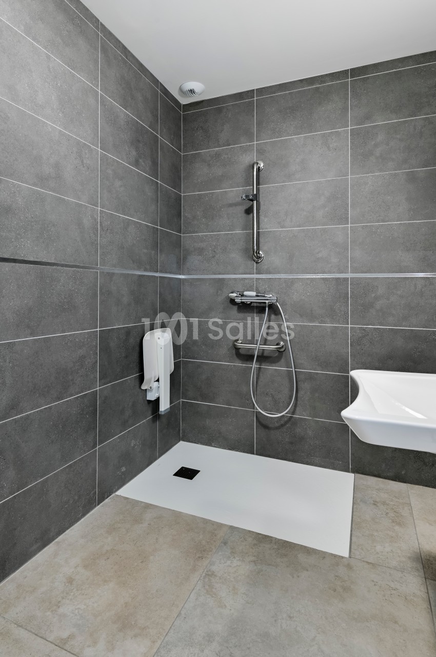 Douche à l'italienne avec carrelage gris, barre d'appui, pommeau réglable et distributeur mural dans une salle de bain.