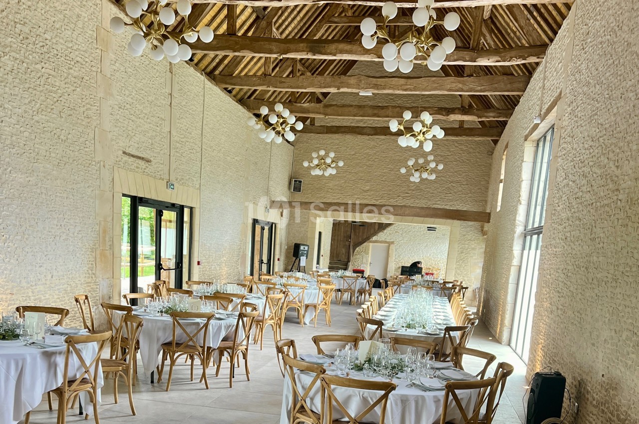 Salle de réception lumineuse avec murs en pierre, tables rondes dressées et poutres apparentes au plafond.