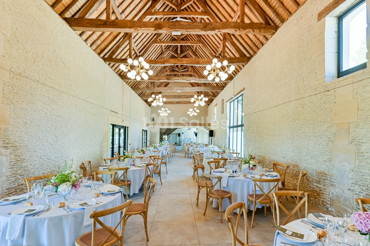 Salle de réception lumineuse avec tables dressées, chaises en bois et poutres apparentes sous un plafond haut.