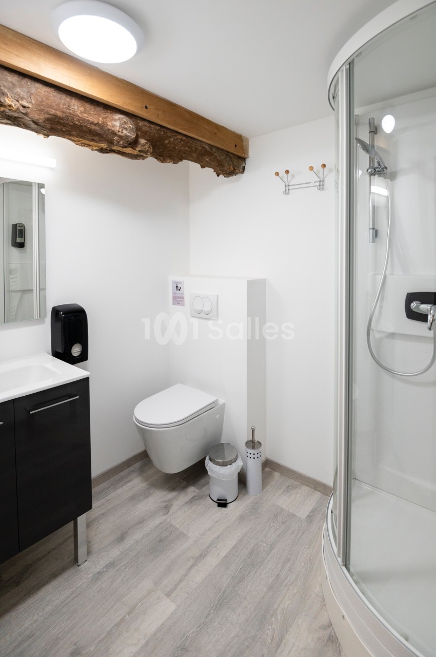Salle de bain moderne avec douche arrondie, toilettes suspendues, lavabo noir et poutre apparente au plafond.