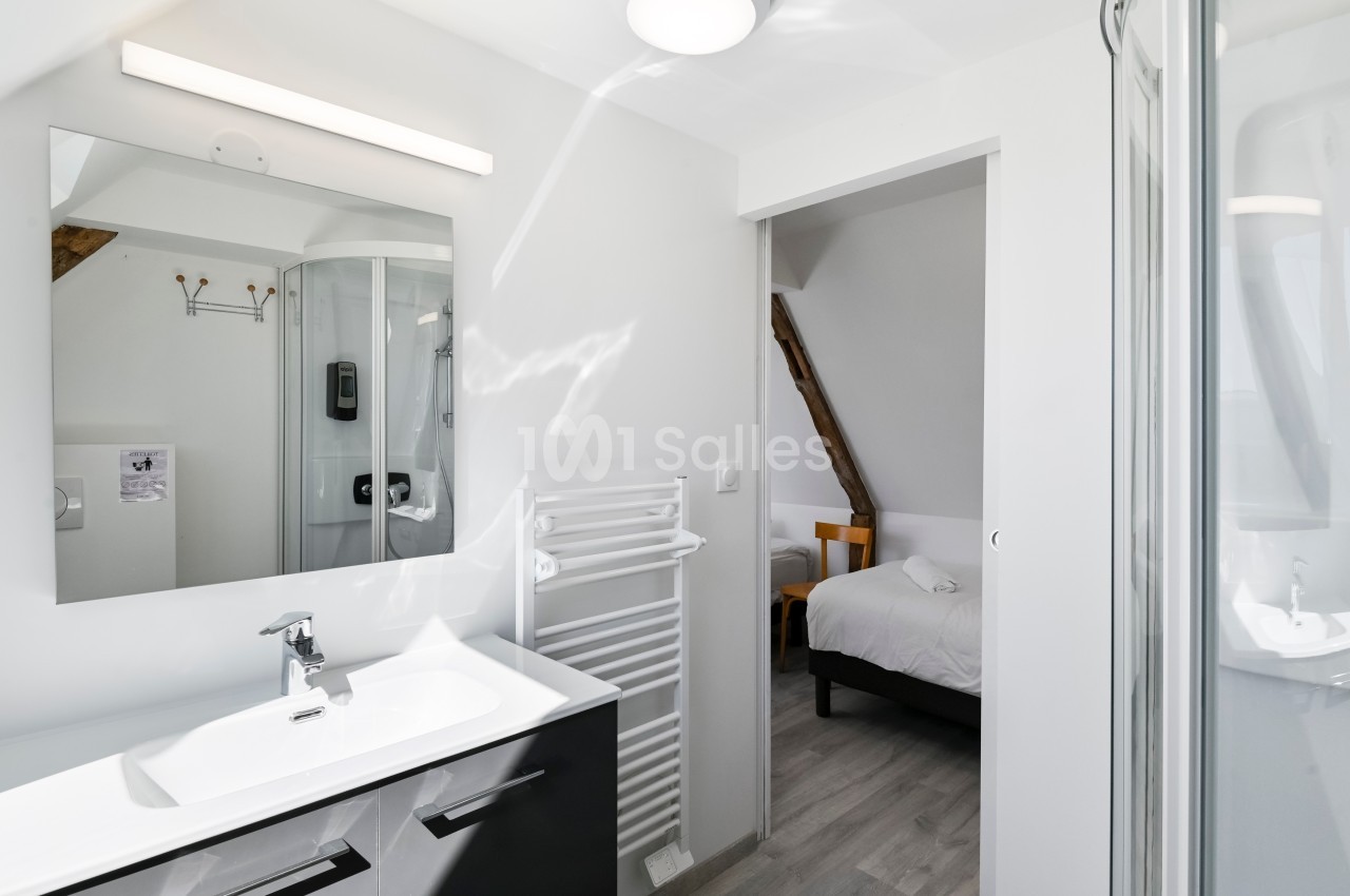 Salle de bain lumineuse avec lavabo, miroir, sèche-serviettes et vue sur une chambre avec lit à travers une porte ouverte.