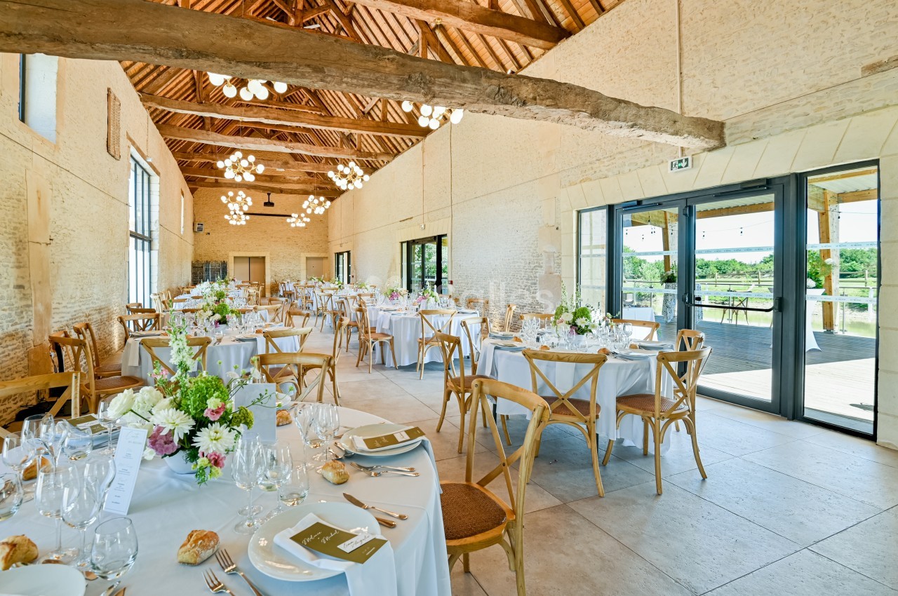 Salle de réception lumineuse avec tables dressées, décorées de fleurs, et vue sur l'extérieur par de grandes baies vitrées.