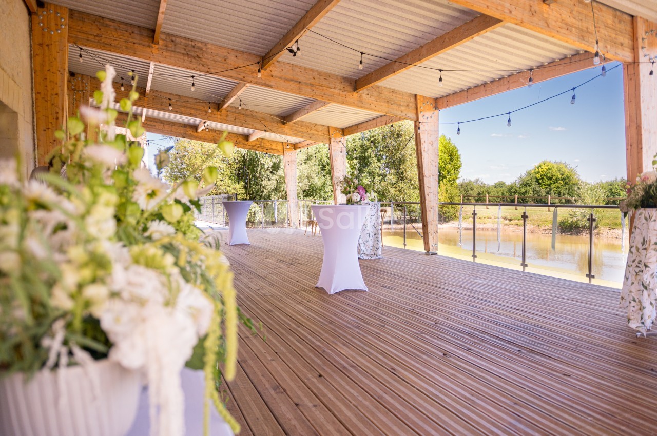 Terrasse en bois couverte avec des tables hautes, vue sur un plan d'eau et décorée de plantes fleuries.
