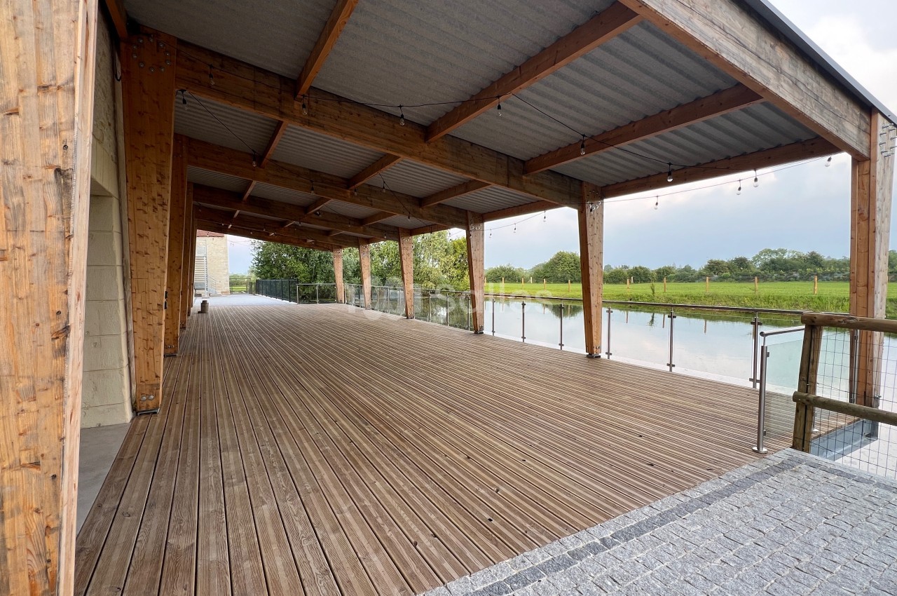 Terrasse en bois couverte avec vue sur un plan d'eau et des espaces verts, entourée de garde-corps en métal et verre.