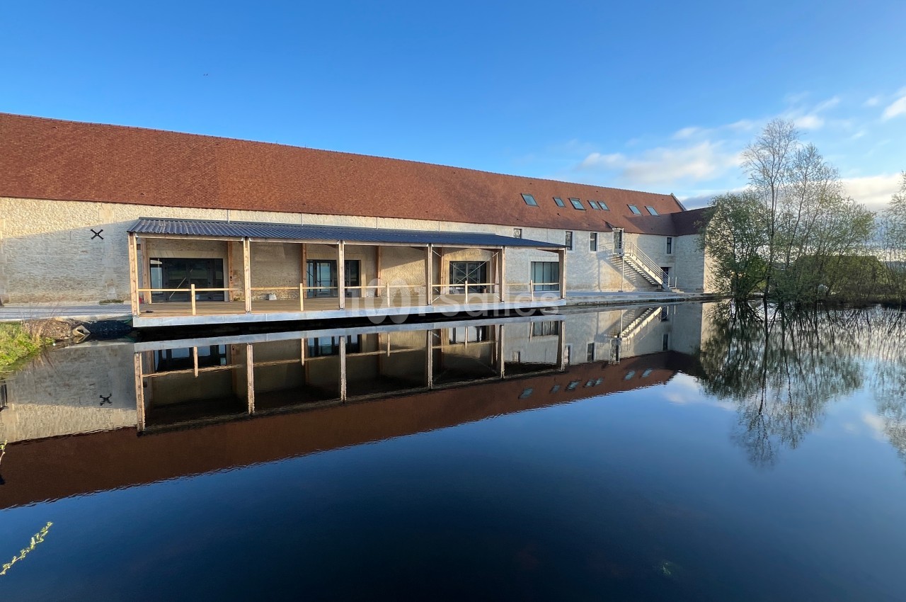 Bâtiment en pierre avec toit rouge, grandes baies vitrées et terrasse en bois, reflété dans un plan d'eau calme.