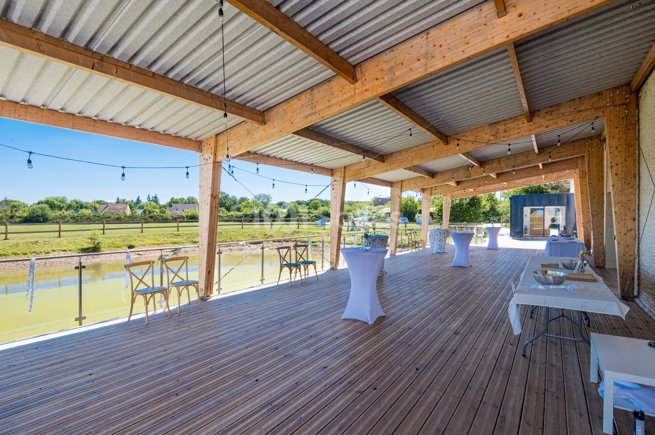 Terrasse couverte en bois avec vue sur un étang, aménagée pour un événement avec tables hautes et chaises.