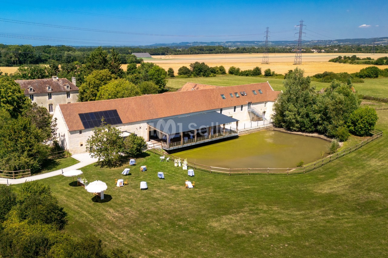 Vue aérienne d'un bâtiment en pierre avec toit rouge, entouré de verdure, d'un étang et de champs à l'arrière-plan.