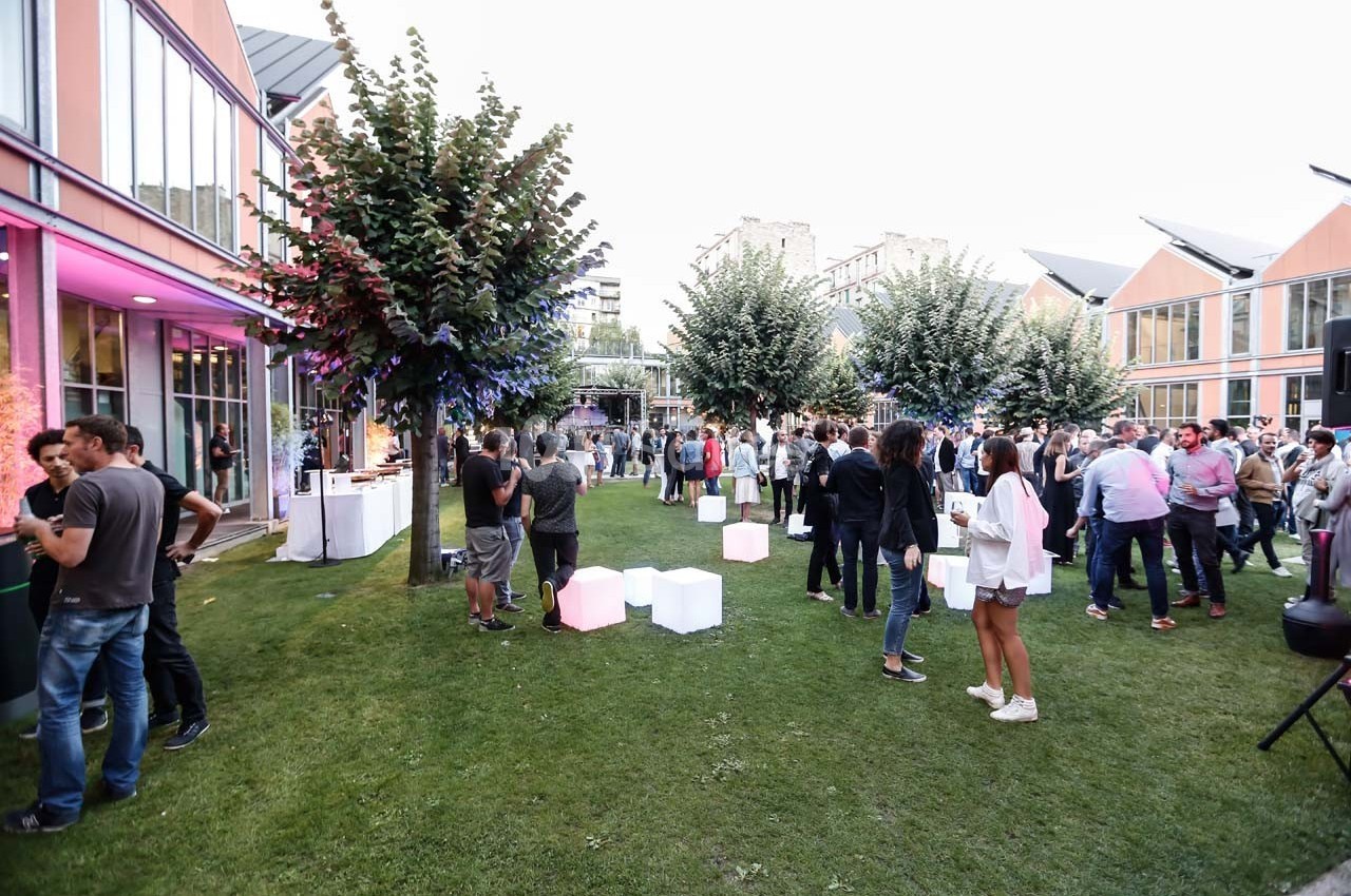 Personnes rassemblées dans un jardin lors d'un événement en plein air, entourées de bâtiments modernes.