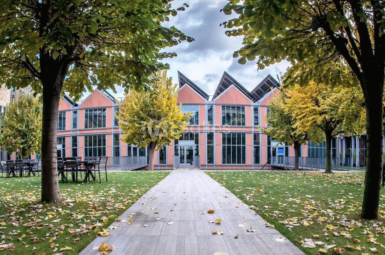 Bâtiment moderne avec façade vitrée, entouré d'arbres aux feuilles jaunies, vu depuis une allée pavée.