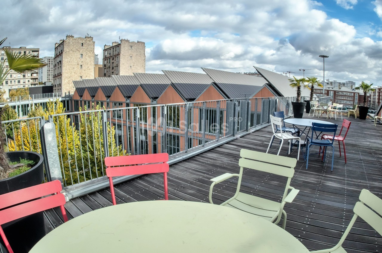 Terrasse en bois avec tables et chaises colorées, vue sur des toits en pente et des bâtiments urbains sous un ciel nuageux.