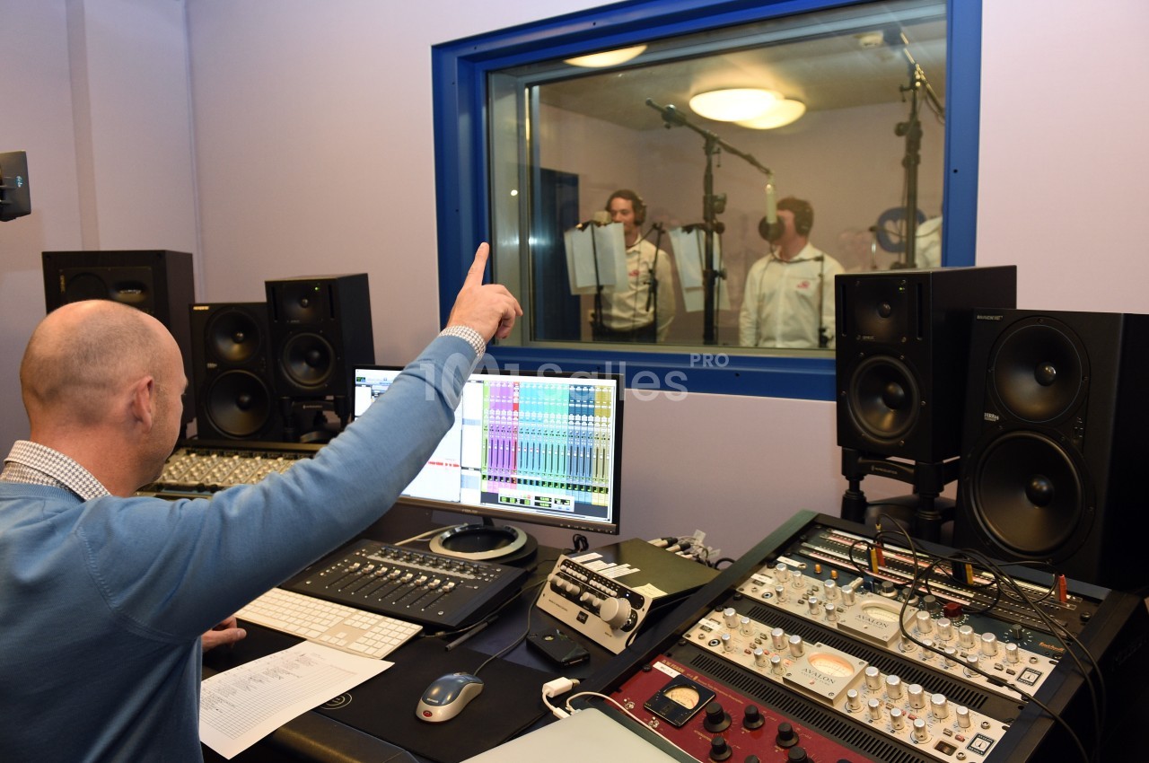Un technicien supervise un enregistrement en studio, avec trois personnes chantant derrière une vitre insonorisée.
