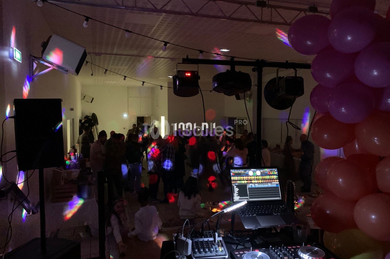 Salle de fête sombre avec des lumières colorées, une table de mixage au premier plan et des invités dansant en arrière-plan.