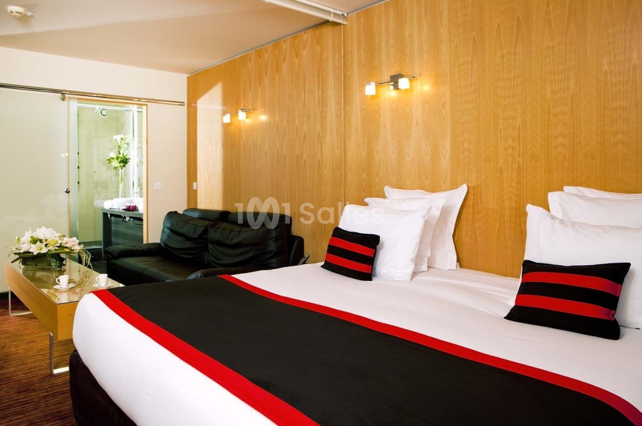 Chambre d'hôtel moderne avec un grand lit, canapé noir, table basse et salle de bain visible à travers une porte vitrée.