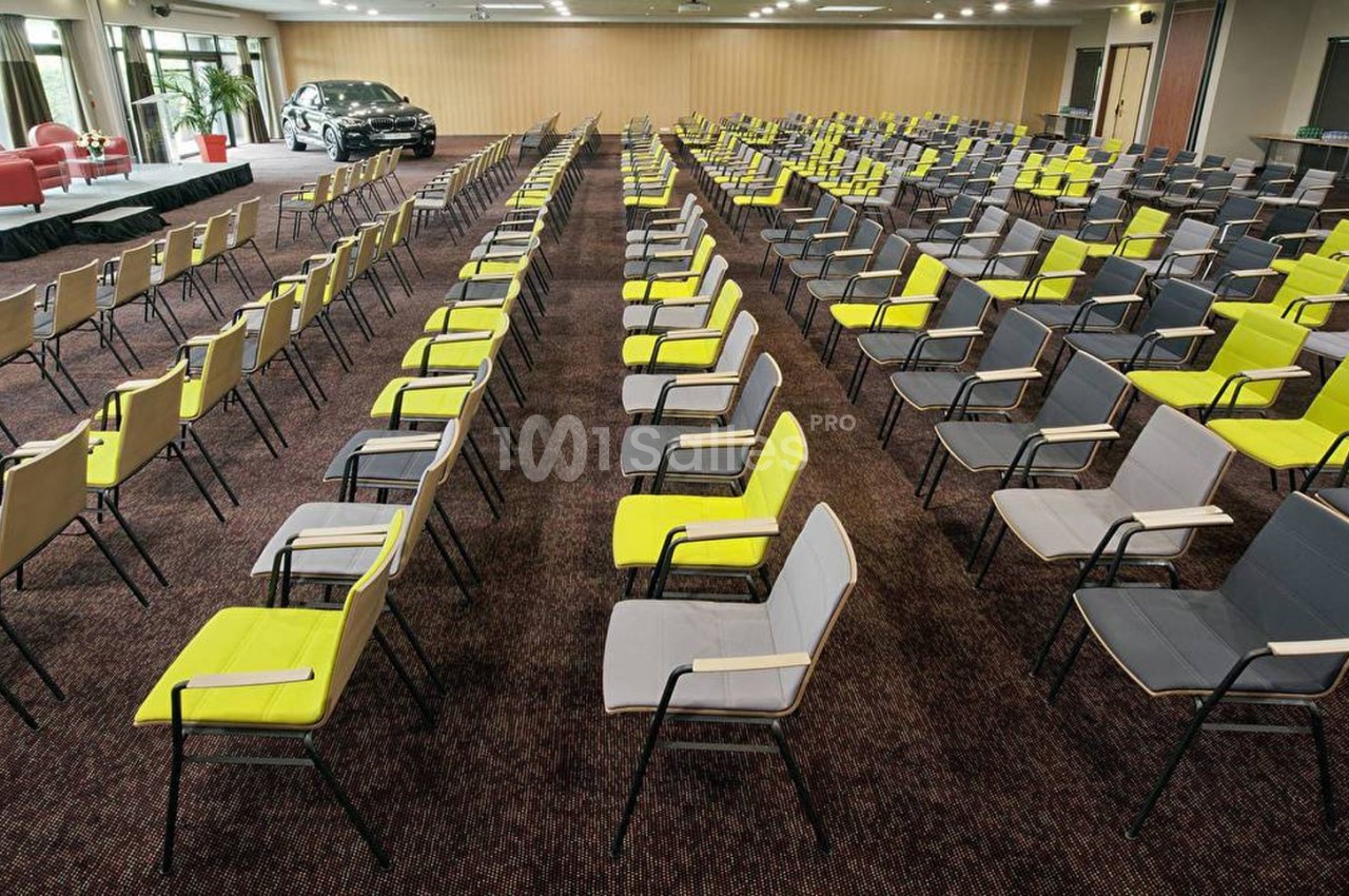 Salle de conférence avec des rangées de chaises grises et jaunes, une scène et une voiture en arrière-plan.