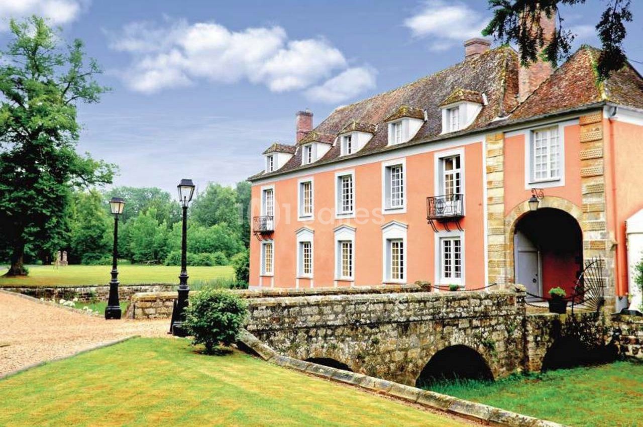 Manoir ancien en pierre avec façade rose, entouré de verdure, traversé par un petit pont en arc.