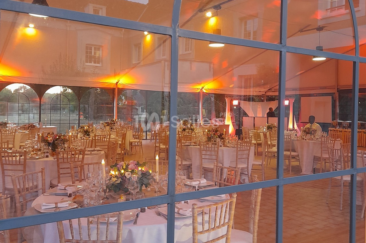 Salle de réception décorée avec des tables dressées, des chaises en bois et un éclairage chaleureux visible à travers une…