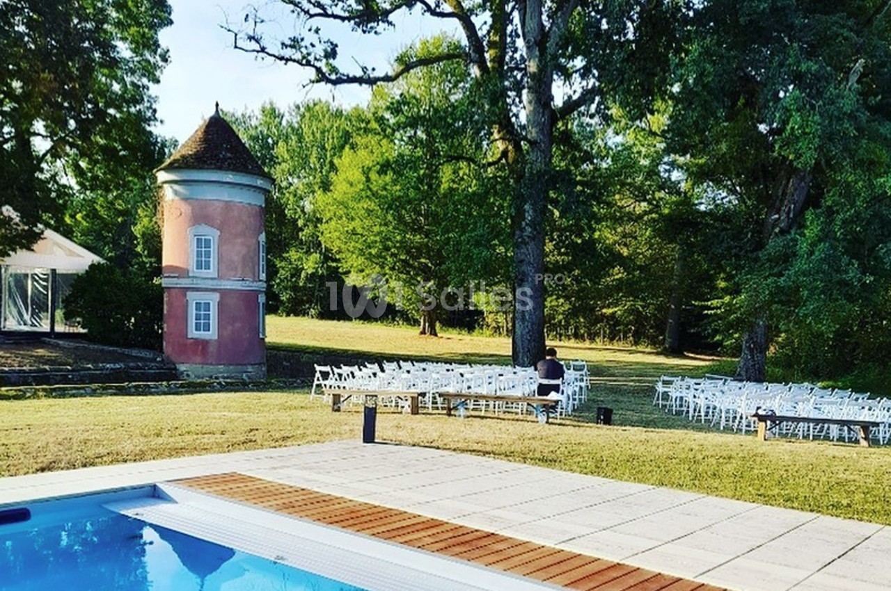 Vue d'une piscine entourée de dalles, avec un petit bâtiment rose, des arbres et des chaises blanches en arrière-plan.