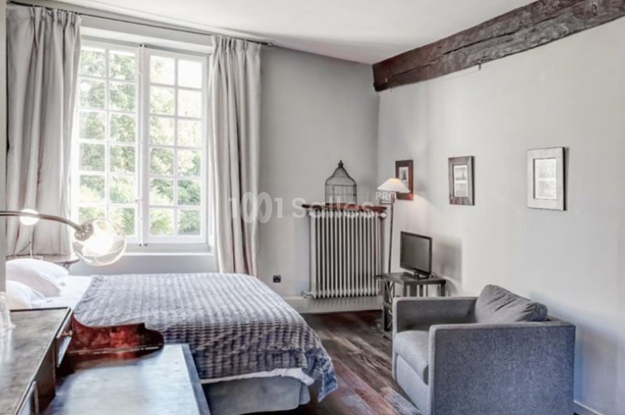 Chambre lumineuse avec lit double, fauteuil gris, bureau en bois, fenêtre à carreaux et poutres apparentes.